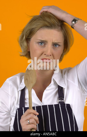 BLOND WOMAN WEARING WHITE BLOUSE ET TABLIER avec cuillère en bois et d'EXPRESSION DE LA DÉTRESSE Banque D'Images