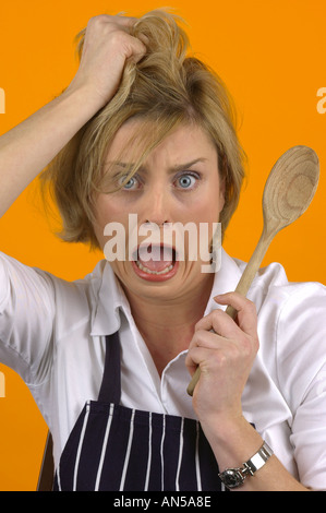 BLOND WOMAN WEARING WHITE BLOUSE ET TABLIER avec cuillère en bois et d'EXPRESSION DE PANIQUE Banque D'Images