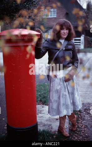 Jeune femme dans une veste en cuir noire et une jupe à rayures gris féminin s'appuyant sur un poste britannique pillar box Banque D'Images