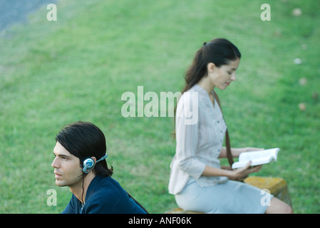 Les gens assis dans un parc, l'homme à l'écoute au casque tout en femme lit book Banque D'Images