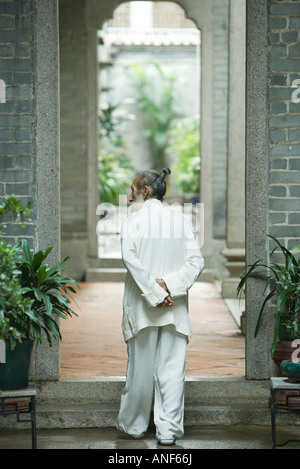 Un homme âgé en chinois traditionnel, en marchant à travers les vêtements avec les mains derrière archway retour Banque D'Images