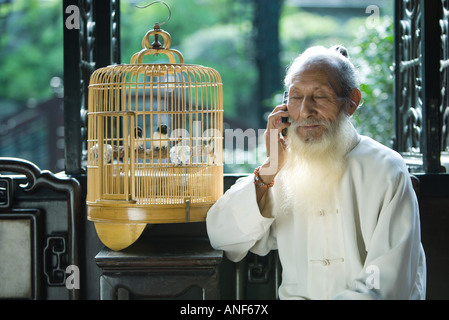 Un homme âgé en vêtements traditionnels chinois cell phone, assis à côté de cage à oiseaux Banque D'Images