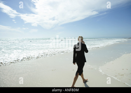 Businesswoman using cell phone, marche pieds nus sur la plage, pleine longueur, vue arrière Banque D'Images