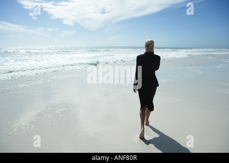 Businesswoman using cell phone, marche pieds nus sur la plage, pleine longueur, vue arrière Banque D'Images