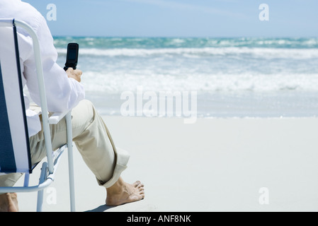 Homme assis en chaise pliante sur plage, holding cell phone, cropped, vue arrière Banque D'Images