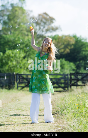 Young woman blowing bubbles in rural field, smiling at camera Banque D'Images