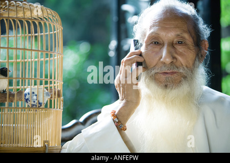 Un homme âgé portant des vêtements traditionnels chinois, en utilisant un téléphone cellulaire, à côté de la cage d'oiseaux Banque D'Images