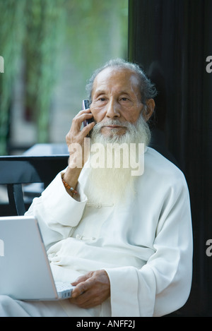 Un homme âgé portant des vêtements traditionnels chinois, à l'aide d'ordinateur portable et téléphone cellulaire Banque D'Images