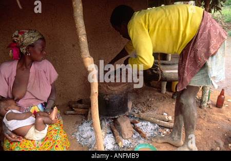 Un homme qui tend la casserole sur un feu ouvert en face de leur maison d'adobe, son épouse assis son bébé de soins infirmiers. Zambézie, Mozambique Banque D'Images