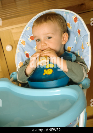 Jeune garçon assis dans une chaise haute de manger un rouleau de saucisse Banque D'Images