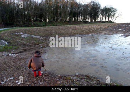 Petit garçon en rouge wellies et un épais manteau pokes une énorme flaque de glace avec un bâton Banque D'Images