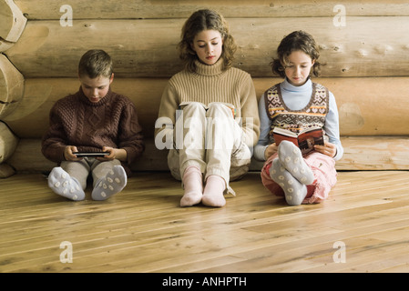 Trois jeunes frères et soeurs assis côte à côte, la lecture et jeux vidéos Banque D'Images