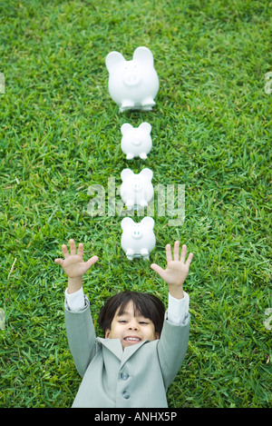 Petit garçon en combinaison complète lying on grass, ligne de Piggy Banks au-dessus de la tête Banque D'Images