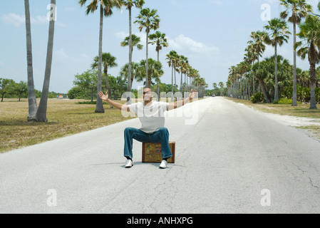 Man sitting on suitcase au milieu de rue, les bras tendus, smiling Banque D'Images