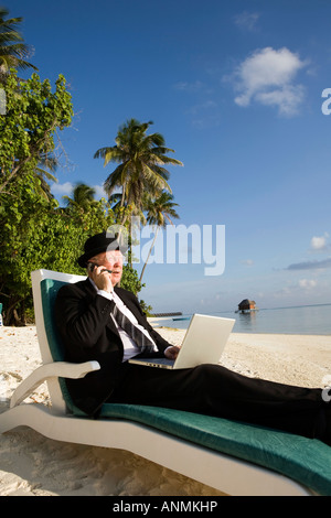 Homme d'affaires en chapeau melon et costume d'affaires travaillant sur un ordinateur portable tout en talking on mobile phone on tropical beach Banque D'Images