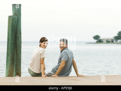 Couple assis sur le bord du quai, smiling over épaules at camera Banque D'Images