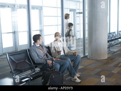 Deux hommes sitting in airport lounge, les voyageurs à pied par en arrière-plan Banque D'Images