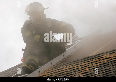 Un pompier sur le toit d'une maison en feu avec un trou de ventilation de toit coupé entourent le pompier avec de la fumée Banque D'Images