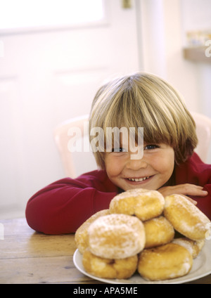 Jeune garçon de s'asseoir à une table de cuisine en souriant à côté d'une assiette de beignets Banque D'Images