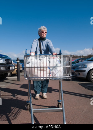 Femme senior retraité en poussant un chariot au moyen d'un parking de supermarché après l'épicerie dans un magasin. Royaume-uni Grande-Bretagne Banque D'Images