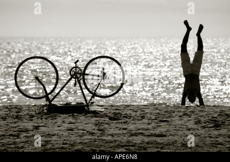 Man doing handstand sur une plage à côté de son vélo tournée vers Banque D'Images