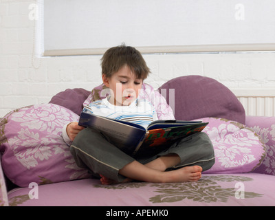 Boy (5-6) reading book on couch in living room Banque D'Images