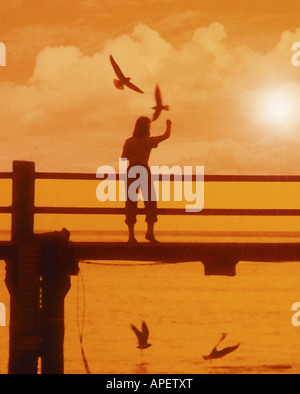 Jeune femme silhouette sur l'alimentation des mouettes planant jetée avec golden fond coucher de soleil. Banque D'Images