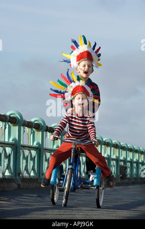 Deux petits garçons en criant des coiffes indiennes rouge comme ils jouent sur un trike sur la voie cyclable sur le front de mer de Brighton Banque D'Images