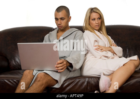 Young mixed race couple sur un canapé portant des peignoirs lui en utilisant un ordinateur portable alors qu'elle a l'air ennuyé Banque D'Images