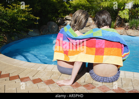 Jeune couple enveloppé dans une serviette de plage, assis à côté d'une piscine extérieure Banque D'Images