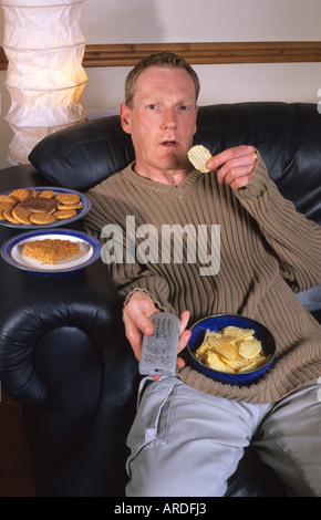 Homme assis sur setee entouré de junkfood utilisant la télévision remote controller Banque D'Images