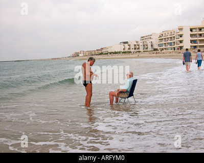 Un homme assis dans une chaise longue dans le surf parle à un homme debout portant les slips Banque D'Images