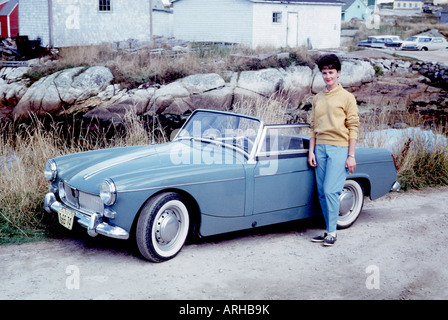 Jolie jeune femme en chandail et pantalon pose par l'océan en face de la voiture de sport MG Midget. Banque D'Images