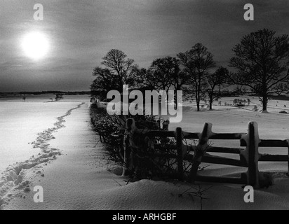 Noir et blanc HIVER PAYSAGE DE NOËL SCÈNE MONTRANT DES TRACES DE PAS DANS LA NEIGE PROFONDE DE TERRAIN AVEC CLÔTURE EN BOIS ARBRES ET UNE ANCIENNE GRANGE Banque D'Images