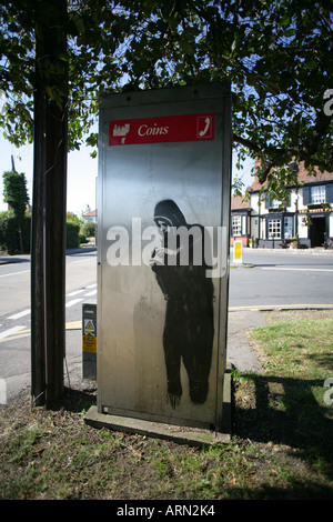 Graffiti au pochoir représentant la figure menaçante d'un jeunes cagoulés sur un téléphone public fort, Essex, Angleterre, Royaume-Uni. Banque D'Images