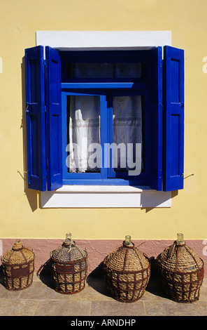 Quatre bonbonnes de verre emballé dans un osier couvrant s'asseoir sous une fenêtre avec des volets bleus lumineux sur l'île de Symi, Grèce Banque D'Images