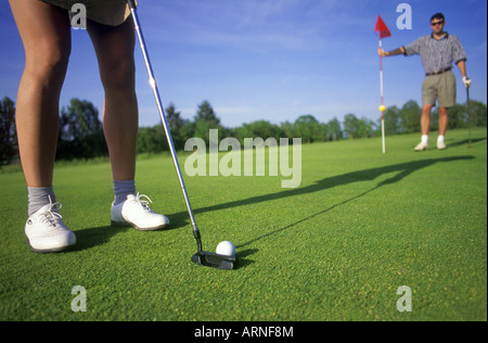 Golfeuse putts sur le green de golf en tant que golfeur mâle aide en soulevant le drapeau du trou, de la Colombie-Britannique, Canada. Banque D'Images