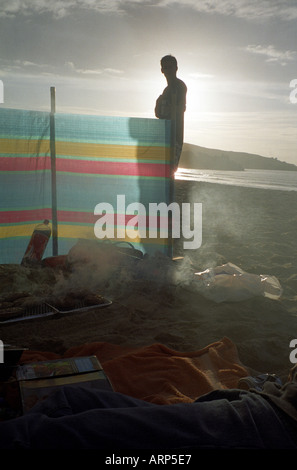 Serviettes et barbecues, rétroéclairés, brise-vent aux couleurs vives et silhouette d'homme sur la plage de Porthmeor, St. Ives, Cornwall, Royaume-Uni. Banque D'Images