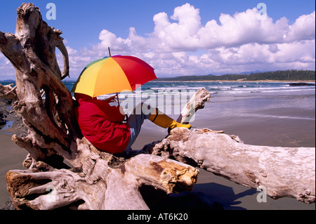 Randonneur avec parapluie posé sur un tronc d'arbre de bois flotté sur un journal West Coast Beach sur l'île de Vancouver, British Columbia Canada Banque D'Images