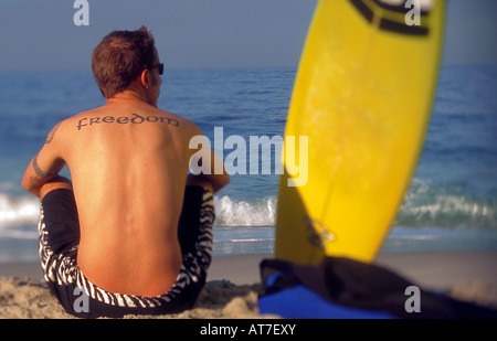 Vue arrière d'un surfeur mâle avec un tatouage lecture liberté assis à côté d'un surf sur la plage Banque D'Images