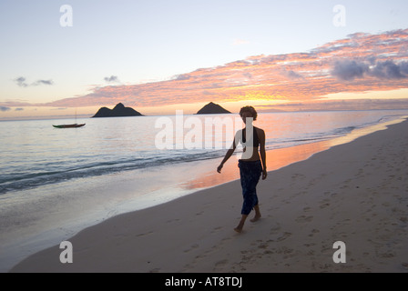 Une femme se promène le long de la plage Lanikai sur le côté au vent d'Oahu avec le Mokulua Islands et un magnifique lever de soleil dans la distance. Banque D'Images