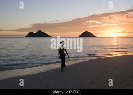 Une femme se promène le long de la plage Lanikai sur le côté au vent d'Oahu avec le Mokulua Islands et un magnifique lever de soleil dans la distance. Banque D'Images