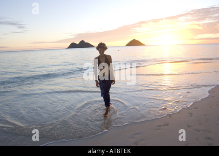 Une femme se promène le long de la plage Lanikai sur le côté au vent d'Oahu avec le Mokulua Islands et un magnifique lever de soleil dans la distance. Banque D'Images