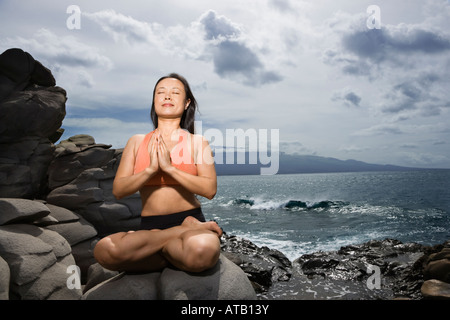 Asian woman sitting on rock par ocean dans lotus posent avec les yeux fermés à Maui Hawaii Banque D'Images