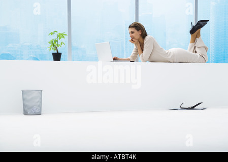 Femme couchée sur le ventre à côté de fenêtre, à l'aide d'ordinateur portable, pleine longueur Banque D'Images