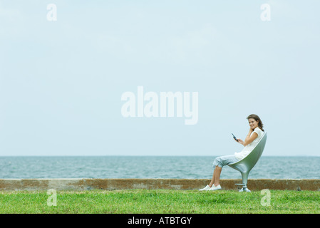 Teenage girl sitting in chair par la mer, holding cell phone Banque D'Images