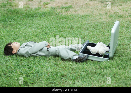 Jeune garçon habillé en costume, couché dans l'herbe à côté de mallette ouverte contenant des ours Banque D'Images