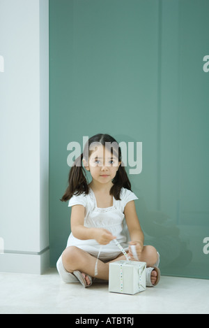 Petite fille assise sur le sol, attachant sur ruban cadeau, looking at camera Banque D'Images