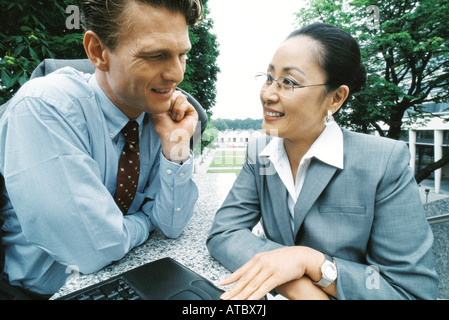 Partenaires d'affaires sitting outdoors, smiling, woman using laptop computer Banque D'Images
