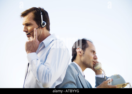 Deux hommes dos à dos, l'un à l'aide du casque, l'autre à l'aide de téléphone fixe, side view Banque D'Images